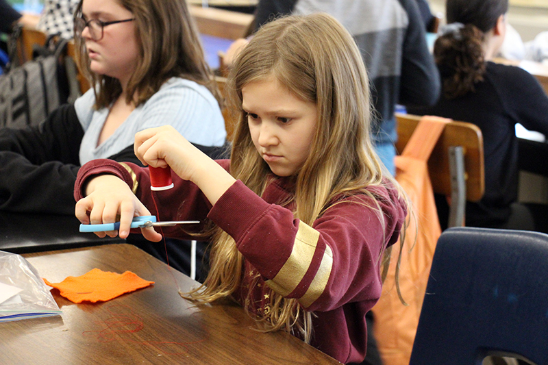 A middle school age girl holds a piece of thread in her left hand and cuts it with scissors.