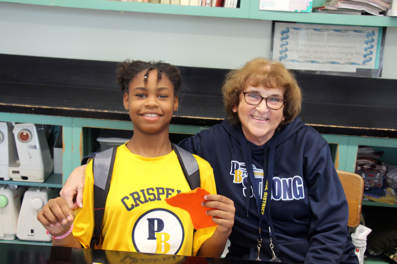 A middle school girl wearing a yellow Crispell Pine Bush tshirt smiles as she sews. A woman sits next to her and smiles too.