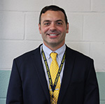 A man in a blue shirt and gold tie and dark suit jacket smiles. He has dark hair.