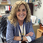 A woman with shoulder-length blonde curly hair sits at a desk with her arms folded on it. She is wearing a light blue jacket and white shirt and is smiling.