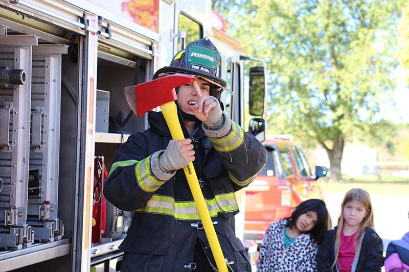 A firefighter dressed in a black coat and a hard firefighter helmet holds up a red and yellow ax and touches the sharp point as elementary students look on.