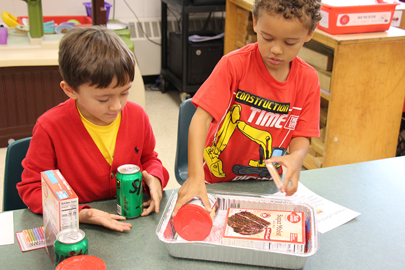 Two kindergarten boys, both dressed in red and yellow, put items in the tin pan.