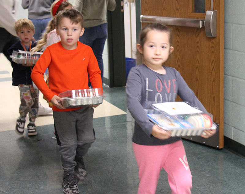 A boy and a girl walk through a hallway, each holding a tin pan with a bunch of goodies inside.