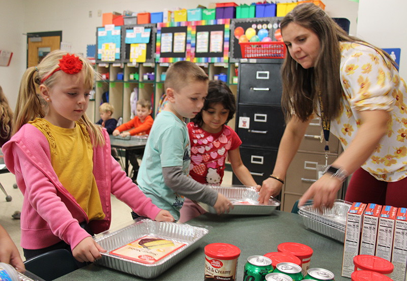 A woman leans over a table to help two kids who are putting items into a tin pan. Another girl, with a big red bow in her hair, has a full pan.