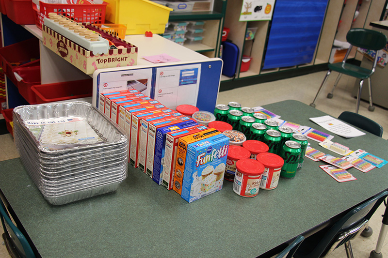 On a table are the following supplies waiting to be boxed and given away: tin 12 baking pans with lids, 12 boxes of cake mix, 12 containers of frosting, 12 cans of Sprite and 12 packages of birthday candles.