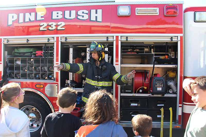 A firefighter points to a kids standing in a group of elementary kids. They are listening to him talk as he stands in front of a red fire truck.