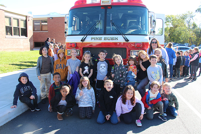 A group of 16 elementary students stand with one adult in front of a big red fire truck.