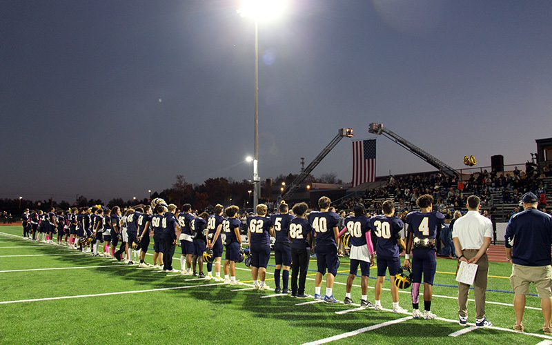 A line of football players, wearing navy blue unifoems with white numbers on their backs, stand at dusk on a field. There is a huge American flag in the back ground.