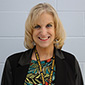 A woman with chin-length blonde hair, wearing a floral shirt and black jacket smiles.