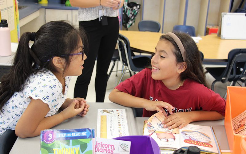 Two second grade girls look at each other and smile as they read books.