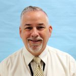 A man with gray hair and a gray goatee, wearing a tan shirt and yellow and blue tie smiles. The background is blue.