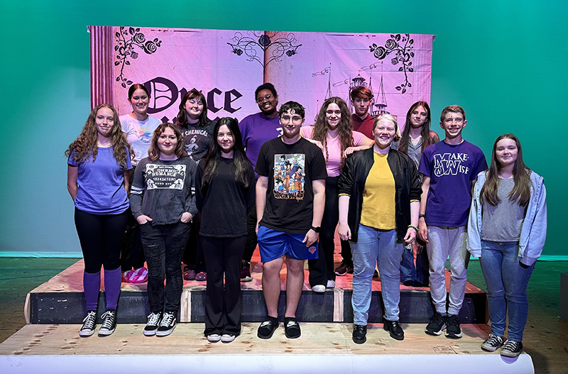 A large group of high school kids standing in three rows in front of a stage background.