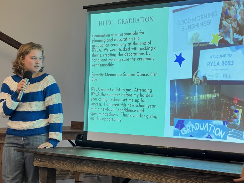 A high school young woman wearing a blue and white striped sweater, presents to a group from what is displayed on a screen.
