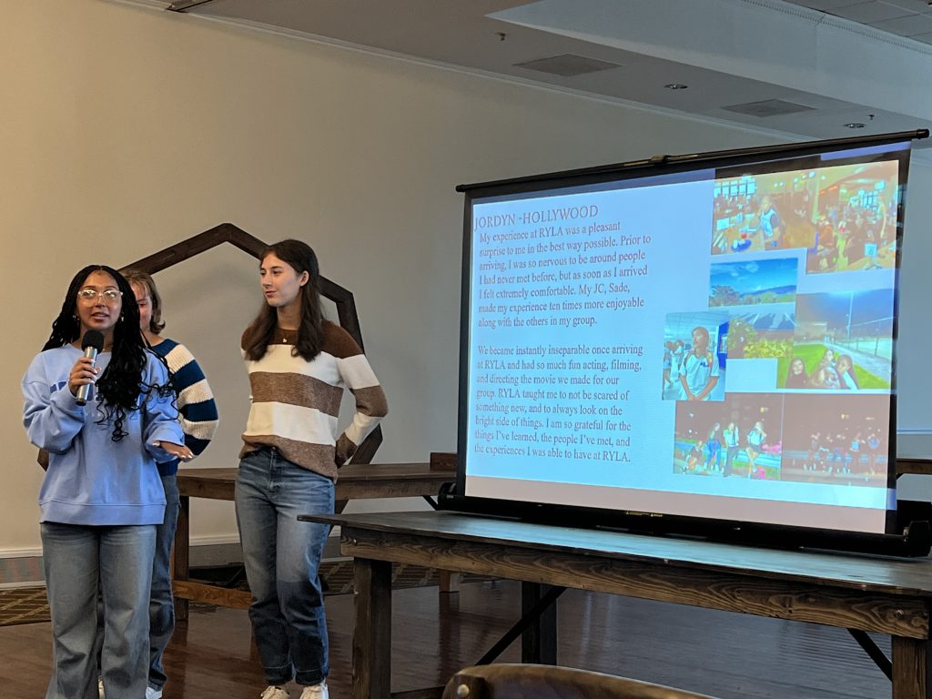 A high school young woman wearing a blue shirt and jeans presents to a group from what is displayed on a screen.
