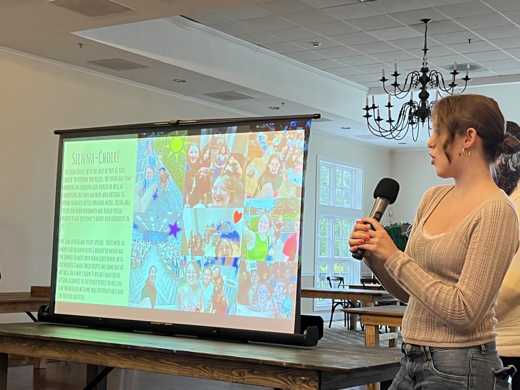 A high school young woman holds a microphone and presents to a group of people from what is displayed on a screen.