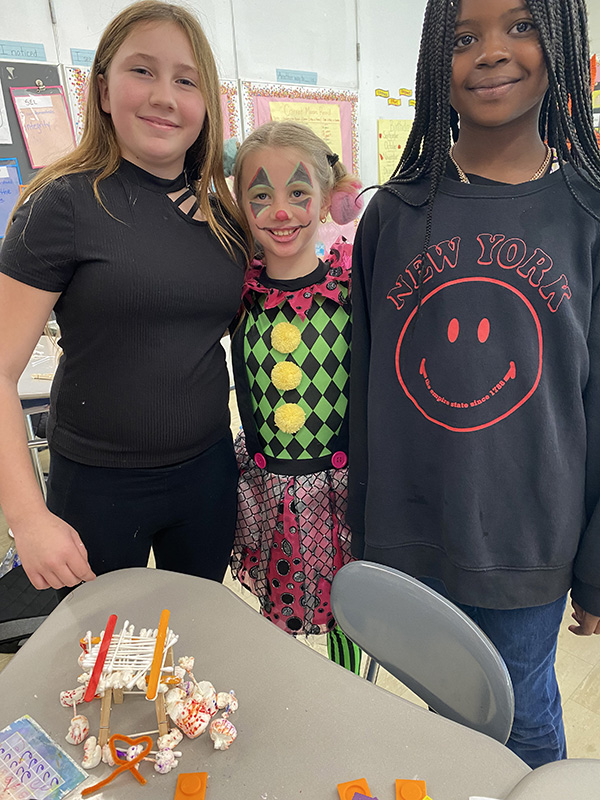 Three girls stand together arm in arm with their homemade bridge on the table in front of them.