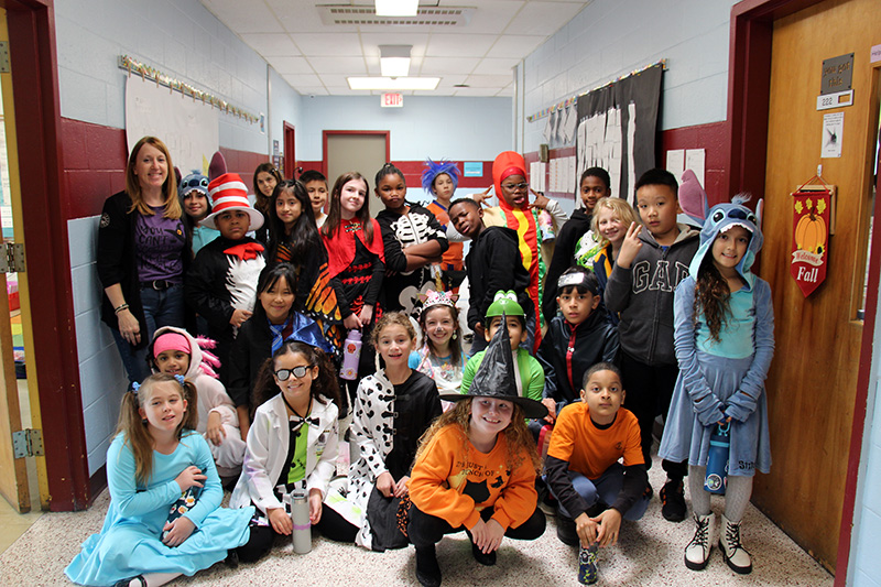 A group of 24 fourth grade students all dressed in Halloween costumes. There costumes are witches, skeletons, cartoon characters, the cat in the hat, a hot dog and many others. There is an adult on the left smiling.