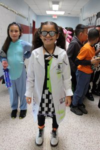 A girl dressed as a chemist - with a lab coat and twirly glasses stands in a hallway smiling.