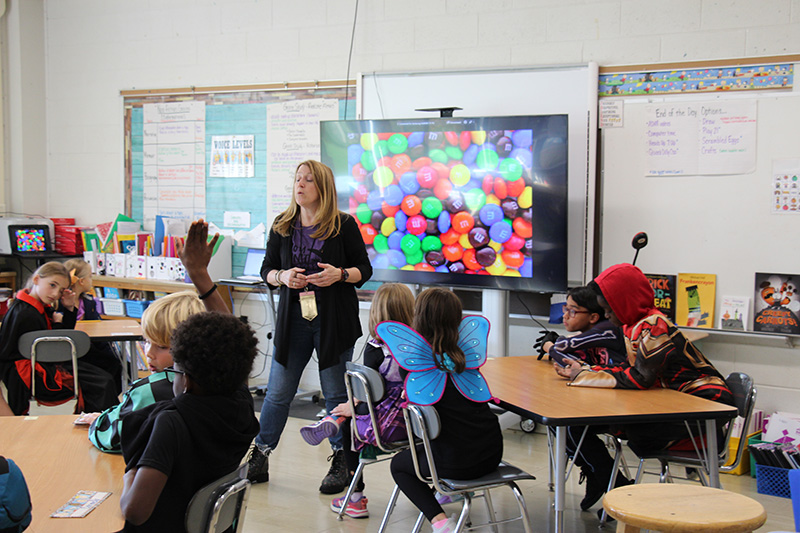 A classroom with students sitting in chairs at tables and a woman in front wiht a screen behind her.