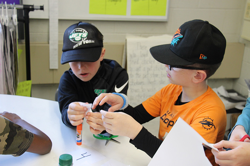 Two boys working together at a table. They are working on creating wings for their paper bat.