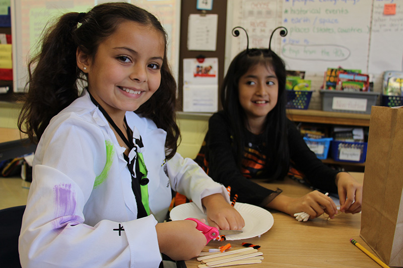 A girl in a white lab coat smiles as she works on building a bridge with different materials. Another girl sits to her right smiling.