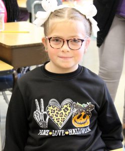 A girl with blonde hair pulled up into ponytails, the holders are ghosts, smiles. She is wearing a shirt that says Peace Love Halloween.