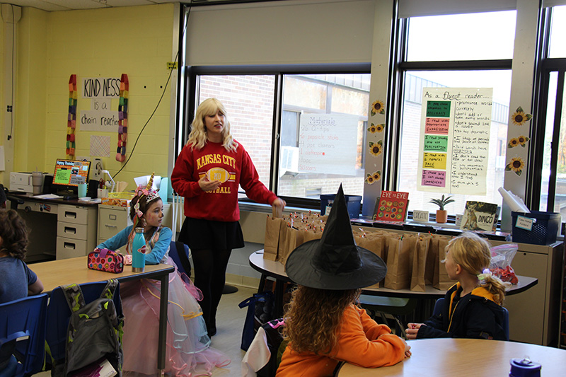 Students sitting at tables while a woman teaches. She is wearing a blonde wig and a black skirt and Kansas City Chiefs sweatshirt on.