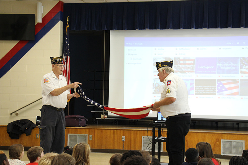 Two men in white shirts, dark pants and blue caps hold a large flag and fold it in front of a group of elementary school kids.