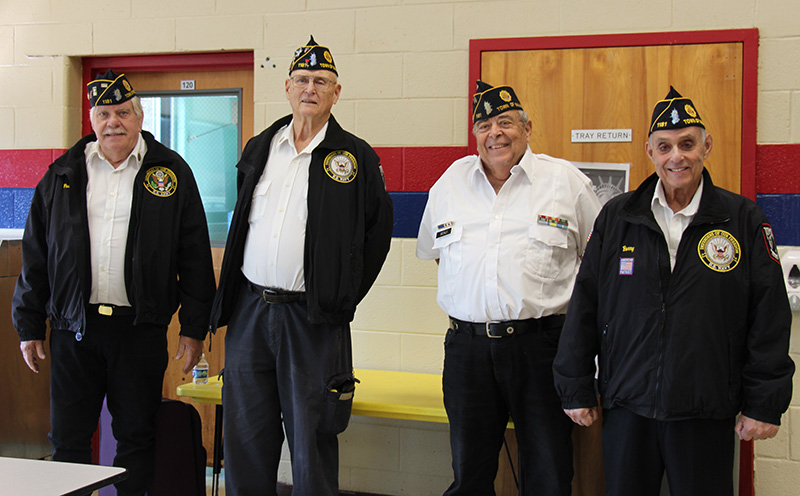 Four men stand together and smile. They are all dressed in white shirts, dark pants and blue caps. Three of them are wearing blue jackets too.