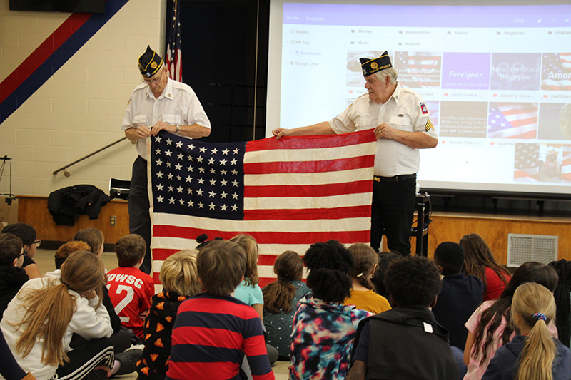 Two en in white shirts and blue caps hold a large American flag in front of a group of students.