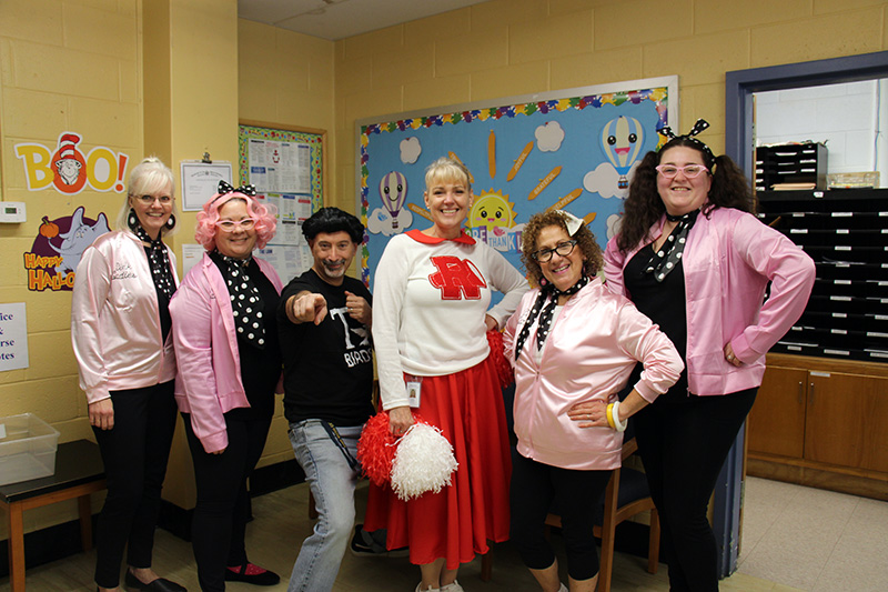Five adults dressed as the characters in the movie Grease. Four pink ladies, two on each side, a man wearing a black wig, black tshirt and jeans, and a woman in red and white cheerleader outfit. all are smiling!