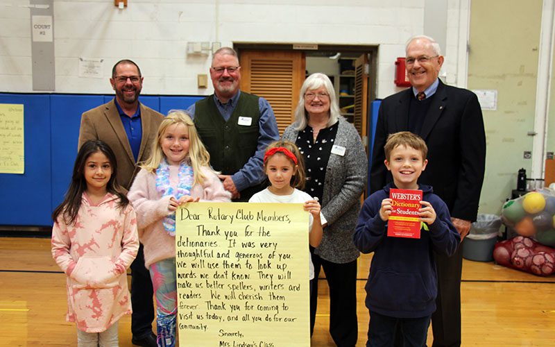 A group of four third-grade kids stand in front while four adults stand in back. The boy on the right holds a red dictionary. The two girls in the center hold a large thank you note and the girl on the left smiles.