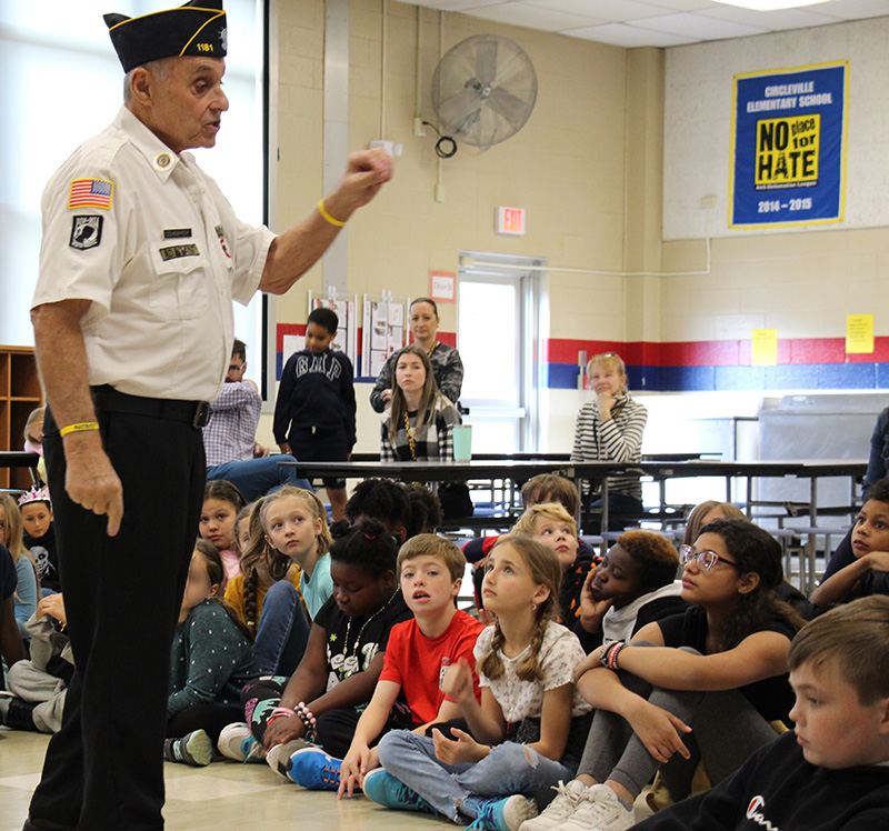 A man in a white shirt with a blue cap talks to a group of fourth grade students who are sitting on the floor.