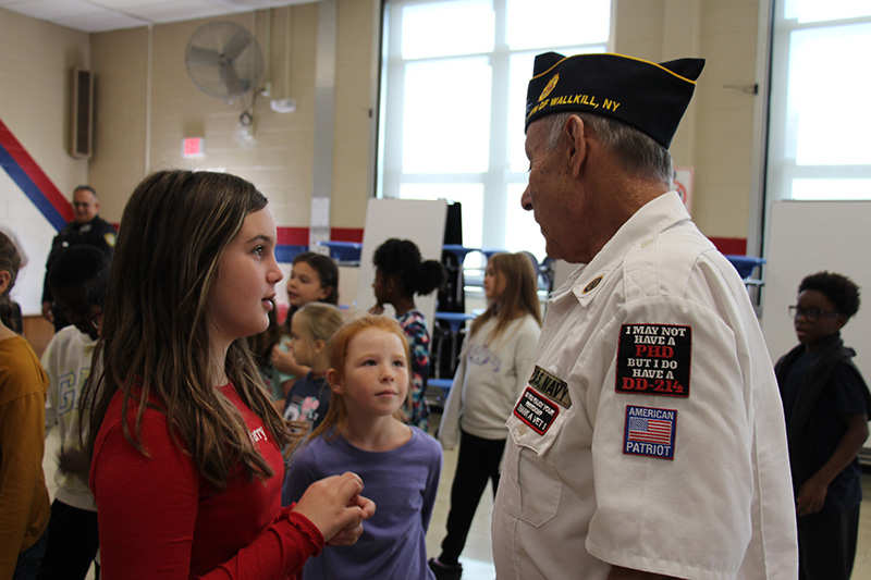 A man in a white shirt and blue cap talks to a fourth-grade student while another one watches and listens.