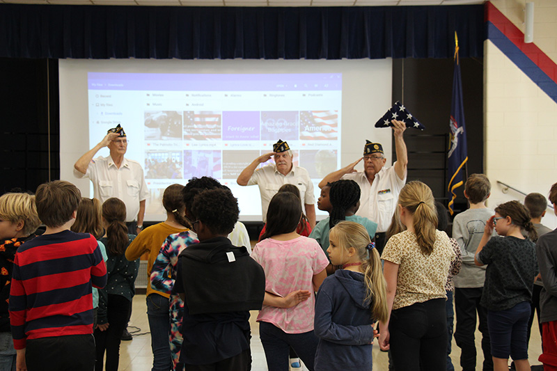 Three men in white shirts and blue caps salute as one holds an American flag, folded into a triangle, high above. A group of fourth-grade students stand too.