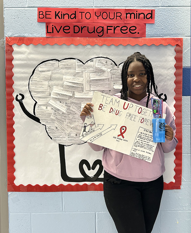 A middle school girl smiles. She has long braids and is wearng a pink shirt and black pants. She is holding a poster she created that says Team up together be drug free forever. Behind her is a bulletin board that says Be Kind to your mind. Live drug free.