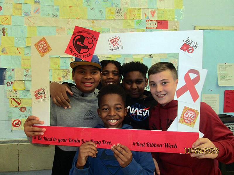A group of five middle school boys smile as they stand inside a photo frame about living drug free.