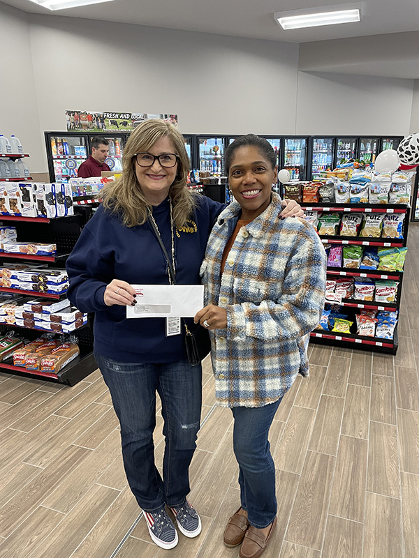 Two women stand inside of a convenience store holding a check.