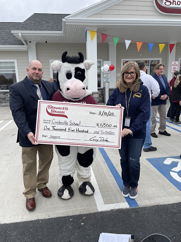 A man on the left, a woman on the right and a person dressed as a cow in the center. The people are holding a large check.