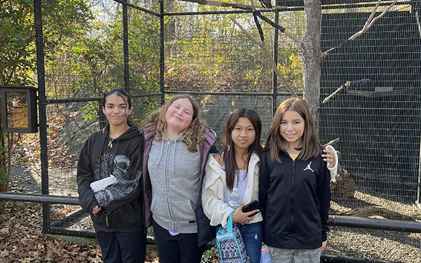 Four sixth-grade girls stand by a fence at a zoo. They are all smiling.