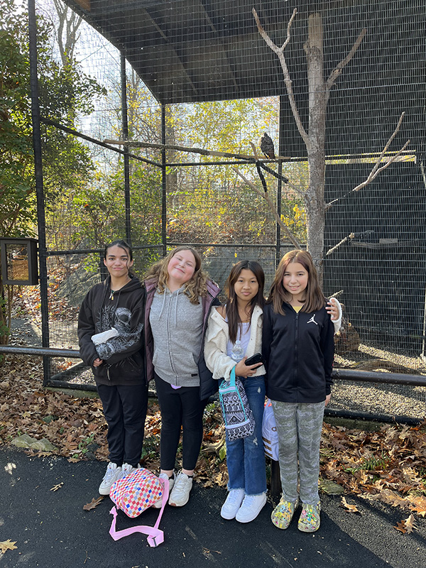 Four sixth-grade girls stand by a fence at a zoo. They are all smiling.