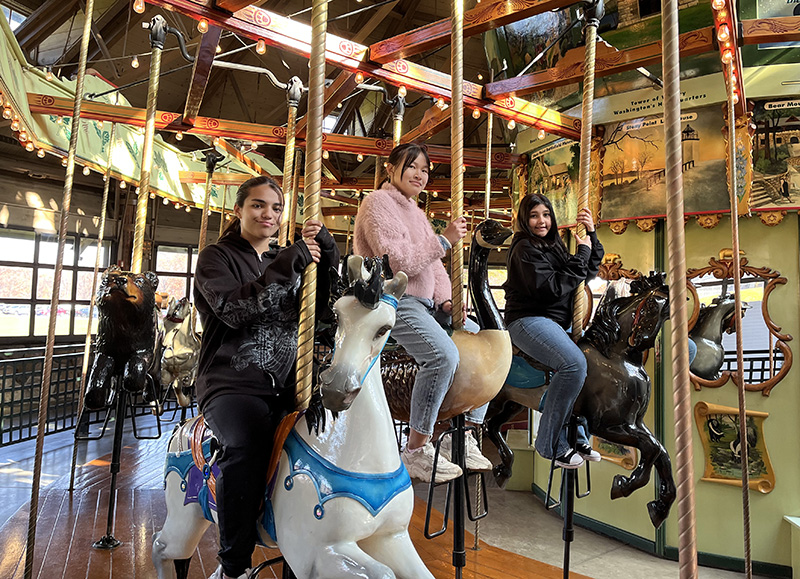 Three sixth-grade kids sit on horses on a carousel. They are smiling.
