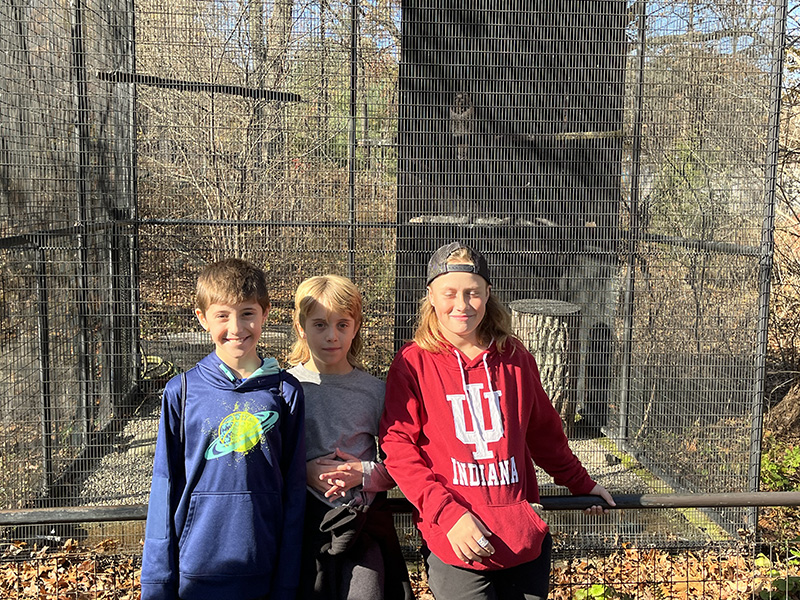 Three sixth-grade boys stand in front of an animal enclosure at a zoo on a sunny fall day.