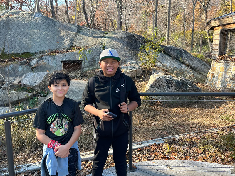 Two sixth-grade students stand in front of an animal enclosure at a zoo on a bright sunny day.