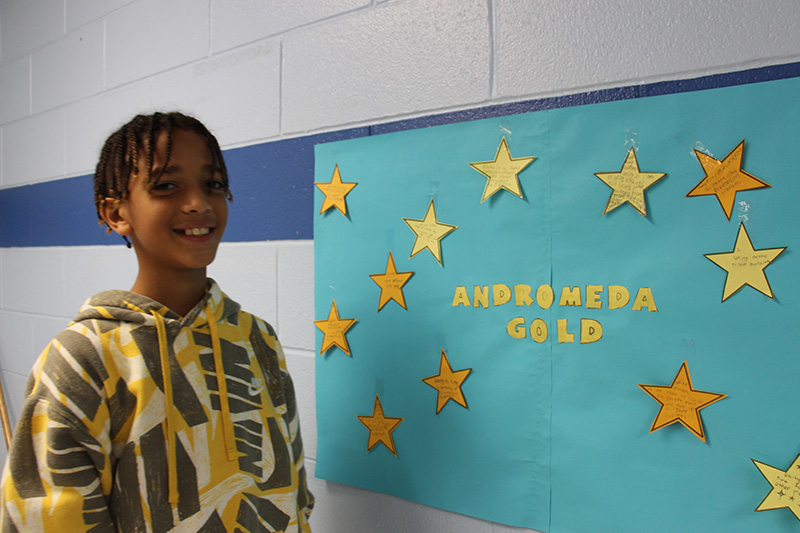 A middle school boy smiles next to a board with a blue background and gold stars on it.