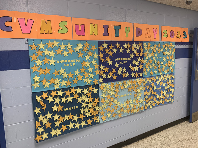 A long hallway wall filled with light and dark blue posters all with gold stars on them. Above it in large colorful letters it says CVMS Unity Day