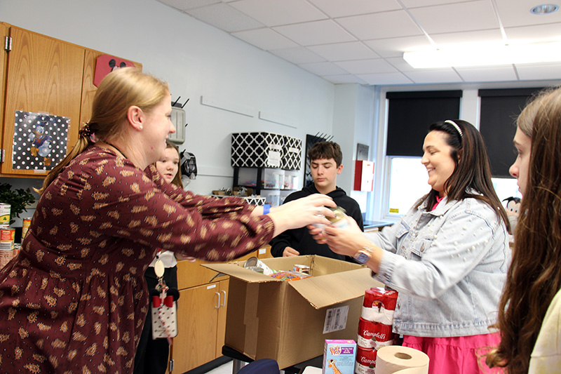 A woman with long blonde hair on the left hands canned food to another woman on the right, with longer dark hair.