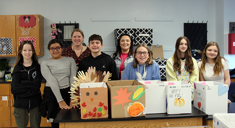 A group of six middle school students stand behind boxes they have filled with food. Behind them are two women. All are smiling.