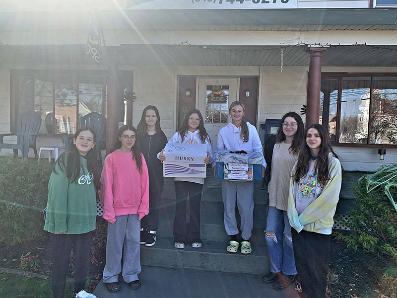 A group of seven middle school girls, two holding boxes, stand on a porch.