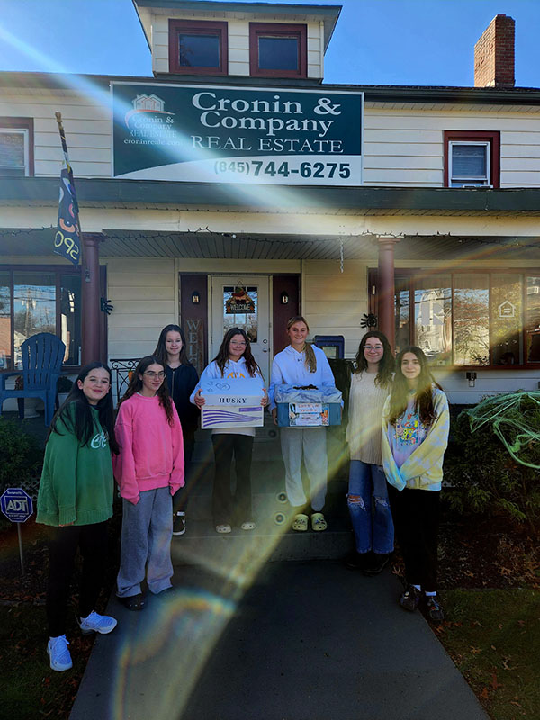 A group of seven middle school girls, two holding boxes, stand on a porch.